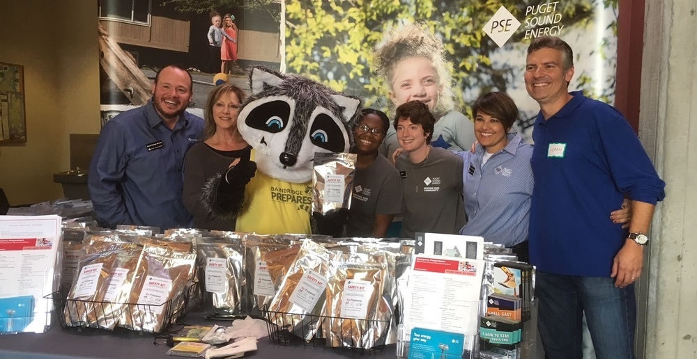 PSE staff with Ready Raccoon at the Expo Day standing behind a table of brochures and packaged food.